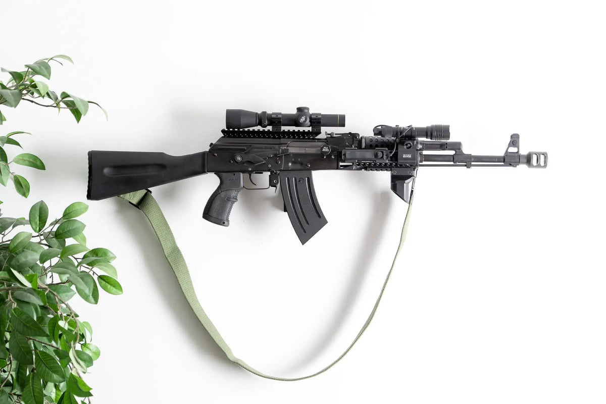 Black rifle with scope on a white wall next to a green plant