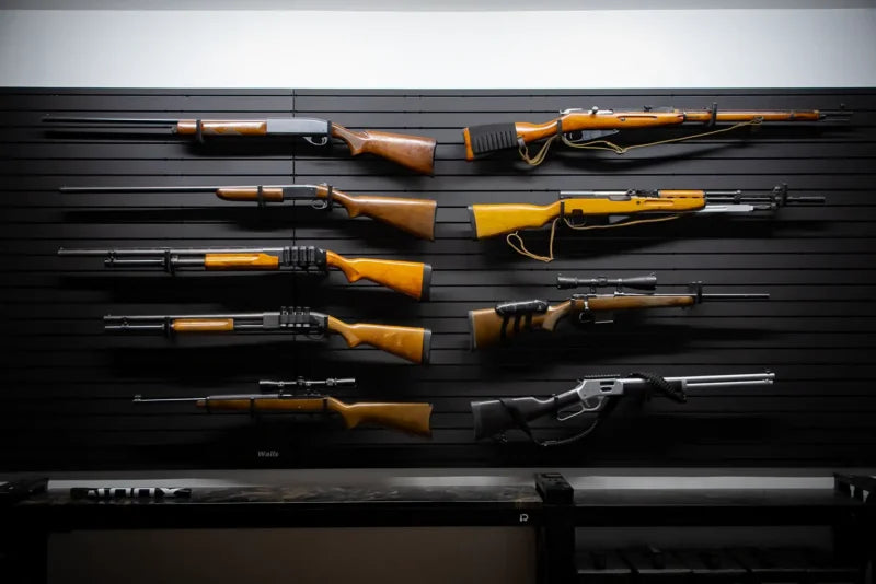 Complete Tactical Walls ModWall panel wall display showing seven rifles and shotguns arranged horizontally across a black paneled wall: bolt-action rifles, pump shotguns, hunting rifles with scopes, and lever-action rifles, all mounted using Tactical Walls MWHRACKRS ModWall rack pairs