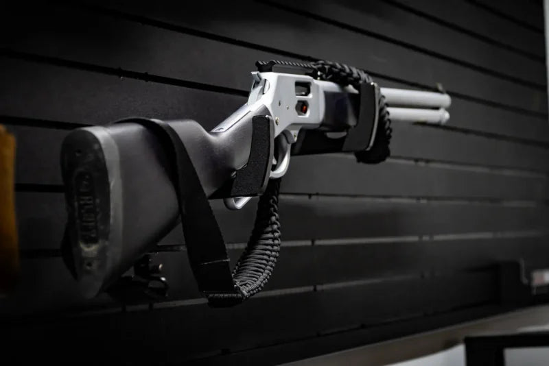 Tactical shotgun with black synthetic stock and paracord sling mounted horizontally on a Tactical Walls MWHRACKRS ModWall rack pair installed on a black ModWall panel, demonstrating shotgun storage configuration