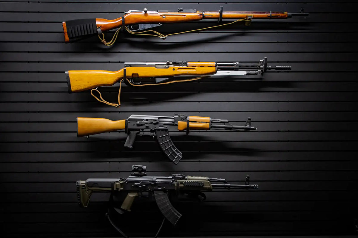 Four rifles displayed horizontally on a black Tactical Walls ModWall panel using multiple MWHRACKRS rack pairs: a Mosin-Nagant bolt-action rifle, an SKS rifle with wooden stock, a wooden-stock AK-47 with 30-round magazine, and a tactical AK variant with green furniture and red dot sight