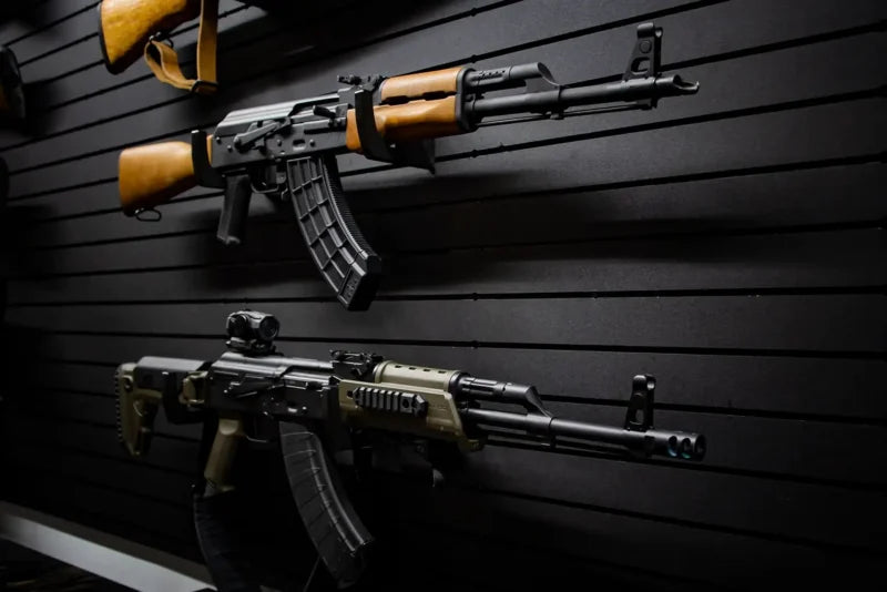 Two AK-style rifles displayed on a black Tactical Walls ModWall panel using MWHRACKRS rack pairs: a traditional wooden-stock AK-47 with 30-round magazine at top and a modern tactical AK variant with tan furniture, red dot sight, and vertical grip below